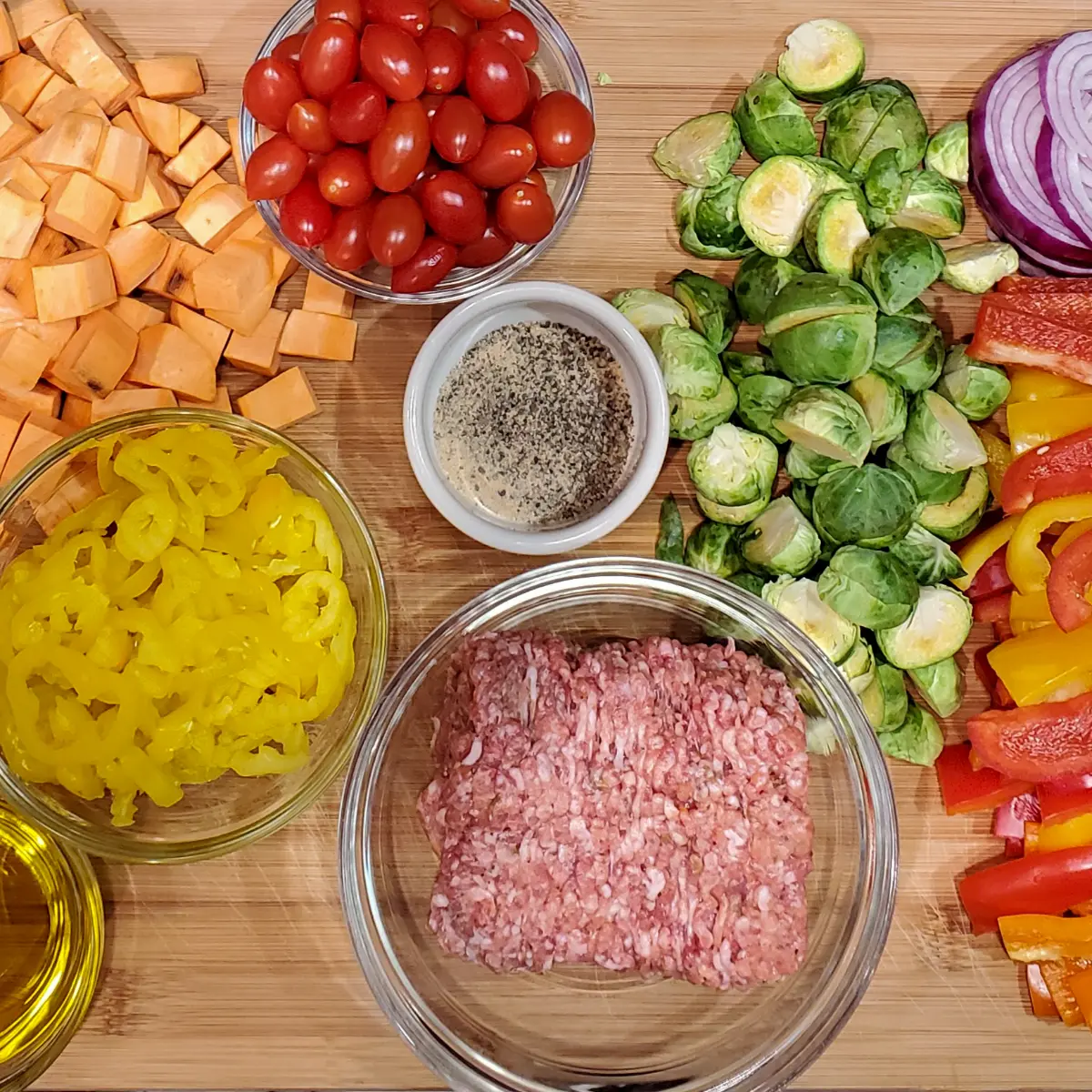 Italian sausage sheet pan recipe from cleveland cooking. All the ingredients are on a large cutting board and are used in the recipe. Italian sausage, olive oil, bell peppers, red onions, sweet potatoes, tomatoes, and banana peppers.