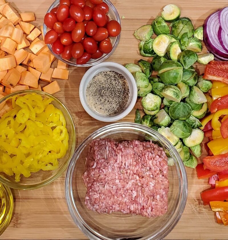 Italian sausage tray bake from cleveland cooking. All the ingredients are on a large cutting board and are used in the recipe. Italian sausage, olive oil, bell peppers, red onions, sweet potatoes, tomatoes, and banana peppers.
