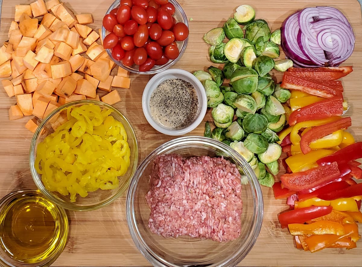 Italian sausage tray bake from cleveland cooking. All the ingredients are on a large cutting board and are used in the recipe. Italian sausage, olive oil, bell peppers, red onions, sweet potatoes, tomatoes, and banana peppers.