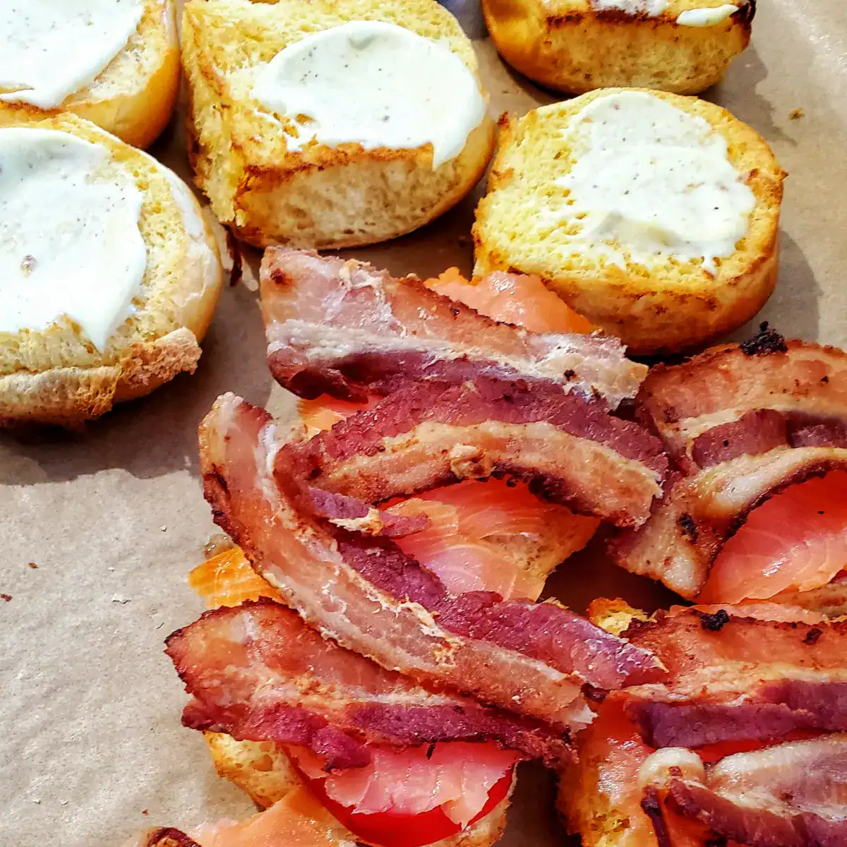 smoked salmon BLT sliders being made. at the top are buns with Dijon mayonnaise and the bottom has the other side of the bun with bacon, lettuce, tomato, and smoked salmon.
