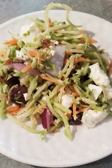 Greek Broccoli Slaw with feta cheese, broccoli slaw, kalamata olives, nuts and a Greek dressing on a white plate.