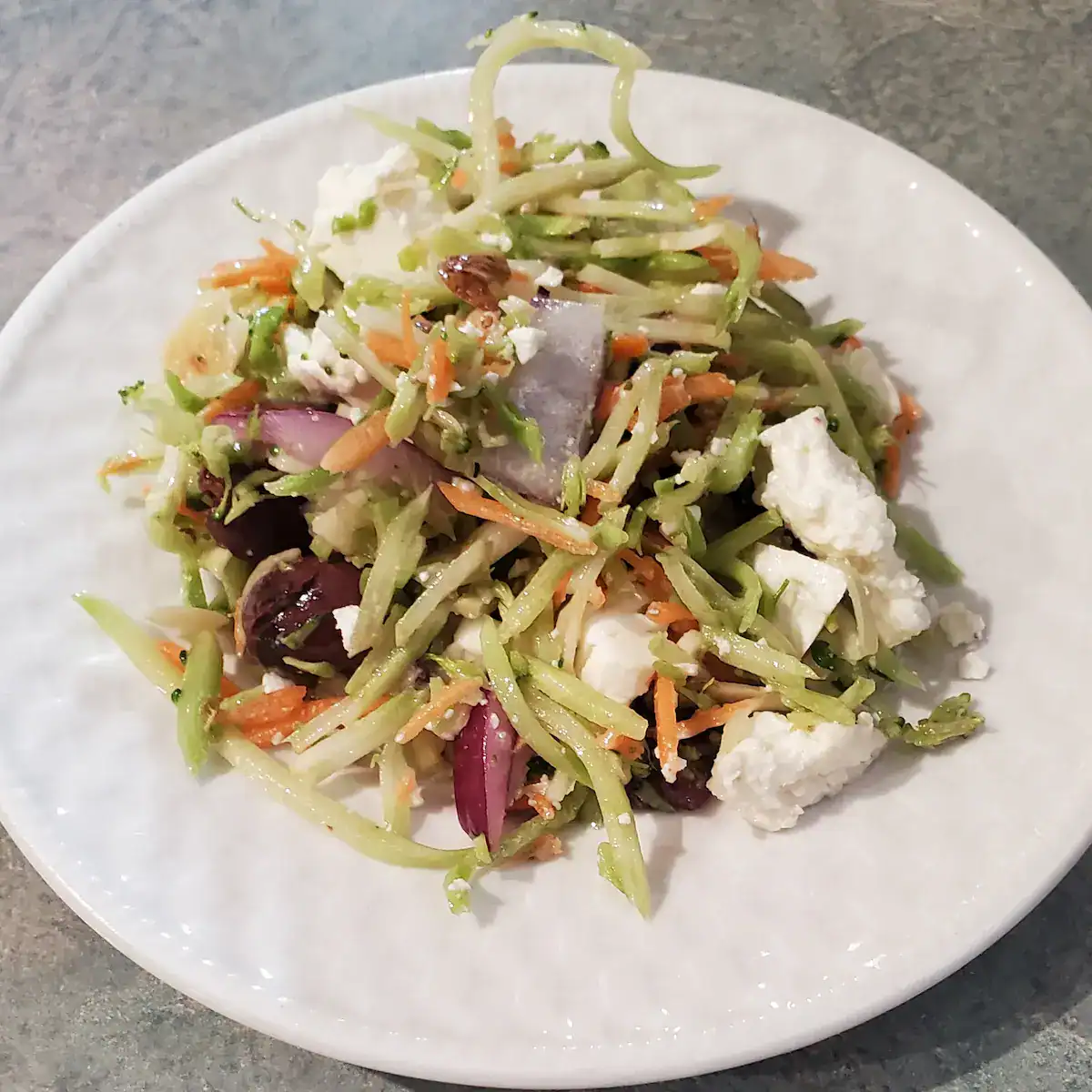 Greek Broccoli Slaw with feta cheese, broccoli slaw, kalamata olives, nuts and a Greek dressing on a white plate.